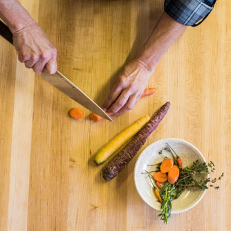 Person cutting carrots on a wooden cutting board with a bowl of chopped vegetables nearby.