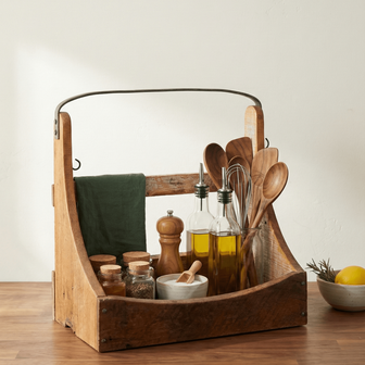 Wooden caddy with kitchen items including bottles, spoons, and a towel on a wooden surface.