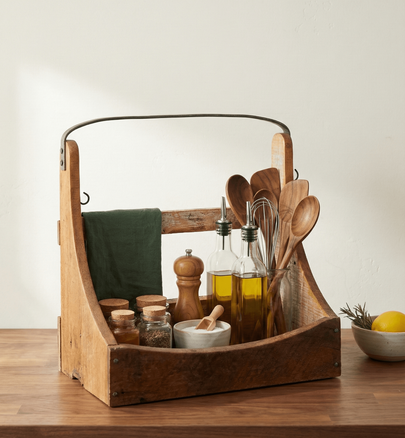 Wooden caddy with kitchen items including bottles, spoons, and a towel on a wooden surface.